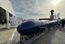 Elta Systems' Blue Whale autonomous submarine. The submarine is seen propped up on display at what appears to be an outdoor expo close to a body of water, slightly visible in the background. The submarine's slender body is painted a deep blue. At least nine individuals are seen scattered across the expo's grounds.