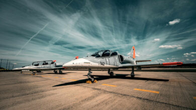 The Hungarian Air Force L-39 Skyfox aircraft is seen stationed on the ground in what appears to be a runway of an air base. The aircraft is painted gray on the upper half of its body, and white on the lower half and on its front. The body number 142 is painted black on its body's left side. The wings and tail's tips are painted a bright red-orange. On its far right, another Skyfox is seen parked. It has the number 144 painted on it.